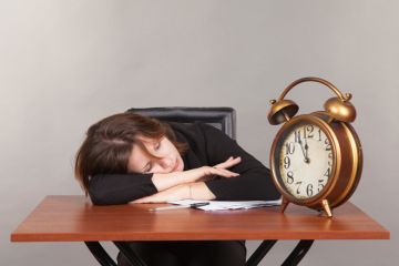 Woman asleep on desk with large alarm clock
