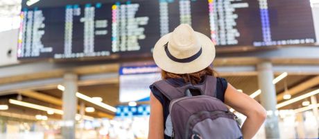 A woman wearing a straw hat and a backpack is standing in front of a large airport display