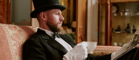 Man wearing formal suit and top hat sitting on sofa reading newspaper
