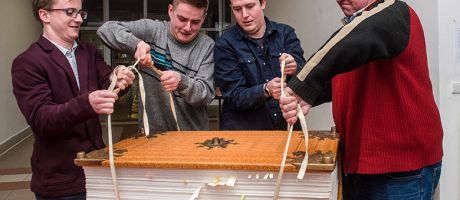 Men struggling to lift a giant book. To illustrate that three major research universities have opted out of new Elsevier deal amid concerns over the cost.