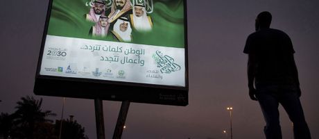 A pedestrian pauses near an illuminated sign showing portraits of the Saudi Arabian Royal family advertising Vision 2030 in Dhahran, Saudi Arabia.
