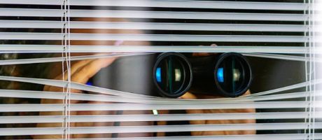 A woman peeking through blinds while holding binoculars, illustrating suspicion A woman peeking through blinds while holding binoculars, illustrating suspicion