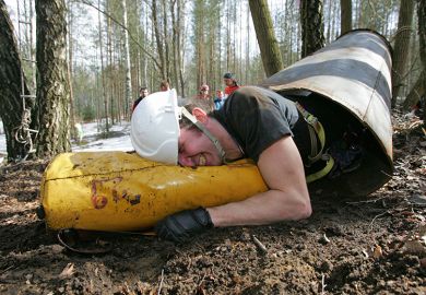 A speleologist crawls with his caving pack through a tunnel during a competition. To illustrate staff feeling forced to publish in a 'narrow' subset of elite journals to boost REF standing.