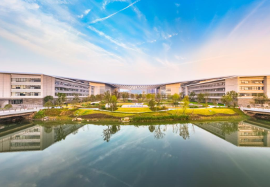 Modern university campus buildings arranged around a landscaped courtyard and reflecting pool, with bridges crossing the water under a bright blue sky.