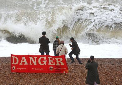 People walk on the beach at high tide, during strong winds in Brighton, United Kingdom, with a sign warning of dangerous seas. To illustrate English universities being ‘unprepared’ as new free speech rules loom.
