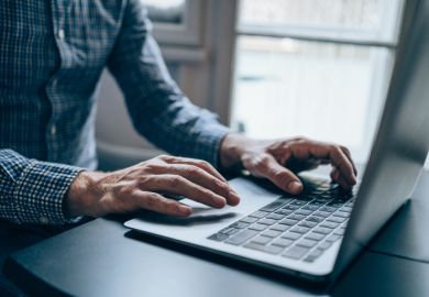 A man wearing a blue chequered shirt, whose face is not visible, typing on a laptop at a desk 