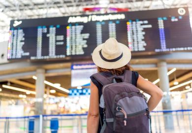 A woman wearing a straw hat and a backpack is standing in front of a large airport display