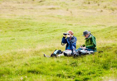 Two senior adults, a man and a woman, sit on the ground in a green meadow in Skye, Scotland