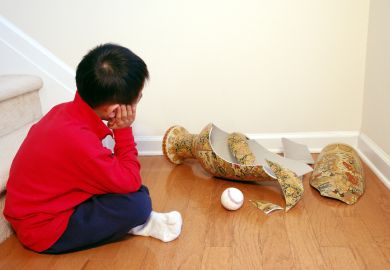 A young boy contemplates a smashed vase, with a ball sitting next to it