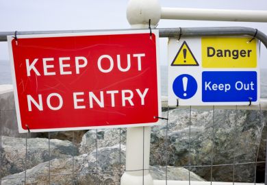 Red and blue 'Keep Out' signs on a fence by the sea, warning of danger ahead.