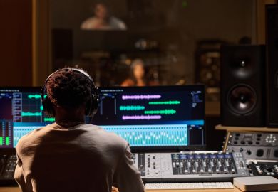 Man wearing headphones operating audio mixing console and computer monitors in professional recording studio, observing musician performing through glass window in background Man wearing headphones operating audio mixing console and computer monitors in professional recording studio, observing musician performing through glass window in background