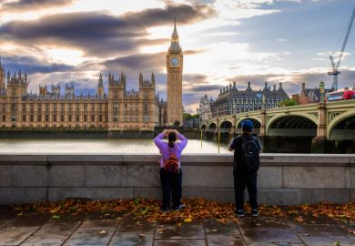 Rear view of a tourist couple taking photos of Big Ben and the Houses of Parliament from the south side of Westminster Bridge on the Thames embankment.