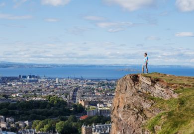 Standing on a rocky precipice, a person overlooks urban scenery and the ocean, under a bright sky Standing on a rocky precipice, a person overlooks urban scenery and the ocean, under a bright sky