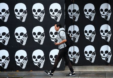A pedestrian, using his cellphone, walks past a boarded establishment showing skeleton heads with cellphones. To illustrate that there is a delay to implementation of the lifelong learning entitlement.
