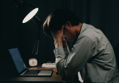 A man working on a laptop holds his head late at night, illustrating overwork