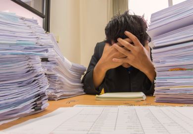 An academic with their head on their desk surrounded by exam scripts