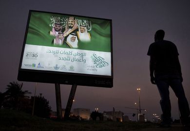 A pedestrian pauses near an illuminated sign showing portraits of the Saudi Arabian Royal family advertising Vision 2030 in Dhahran, Saudi Arabia.