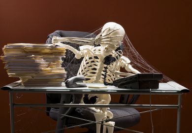 Skeleton sitting at desk on telephone, with webs and stacks of paperwork. To illustrate that colleges are in limbo amid ‘two-year wait’ for degree-awarding powers.