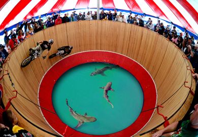 Performers ride motorbikes on a wall of death, with sharks in a pool below. To illustrate that uncertainty is driving Australian universities up the wall. Performers ride motorbikes on a wall of death, with sharks in a pool below. To illustrate that uncertainty is driving Australian universities up the wall.
