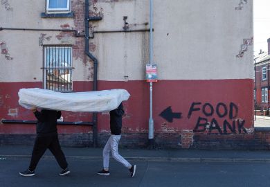 Two men carry a mattress over their heads past a terraced house, the walls of which bare graffiti reading "food bank" to illustrate Cost of living crisis bites for PhDs on ‘inadequate’ stipends