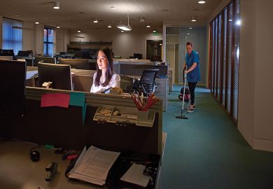 a young woman has stayed behind to get her work done in a large open plan office . A cleaner is vacuuming behind her .to illustrate ‘Illegal’ terms and conditions add to academic overwork crisis