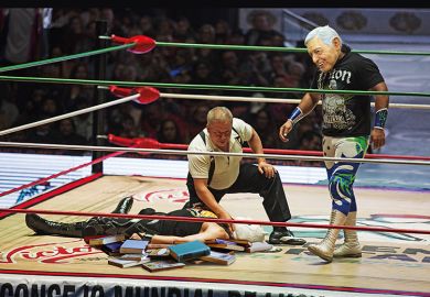 In a wrestling ring filled with books, one man lies prone and is being counted out while his masked opponent prowls