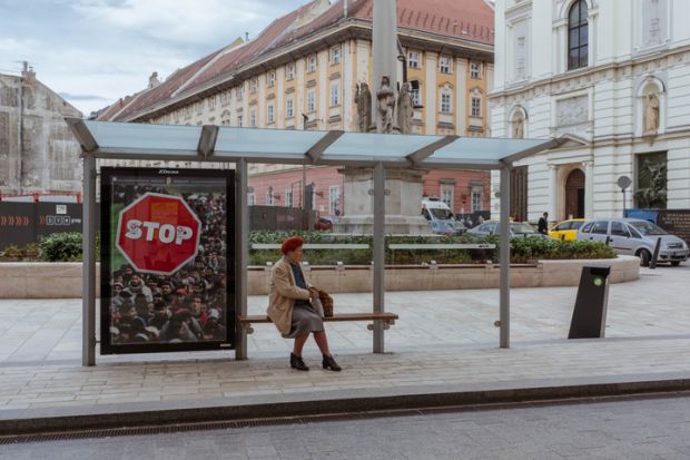 Budapest, Hungary - April 10, 2018 Elderly woman sitting in a bus stop with anti-immigration poster behind her
