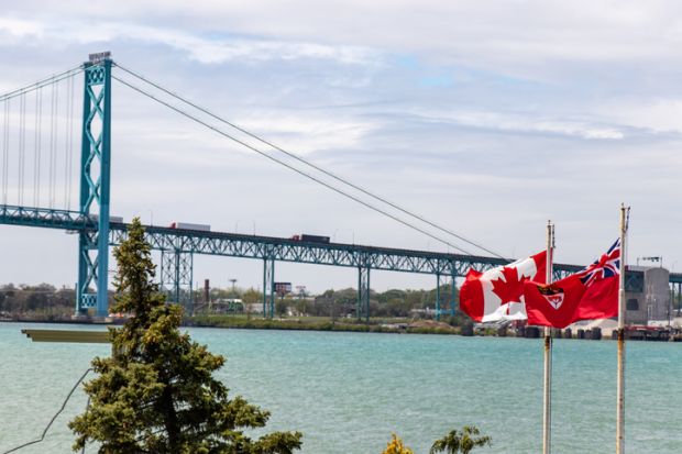 Canada and Ontario flags seen waving in-front of the the US-Canada border, Ambassador Bridge crossing on sunny day.