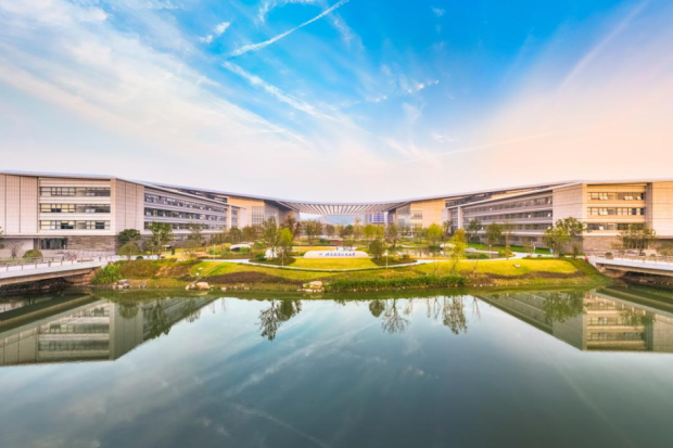 Modern university campus buildings arranged around a landscaped courtyard and reflecting pool, with bridges crossing the water under a bright blue sky.
