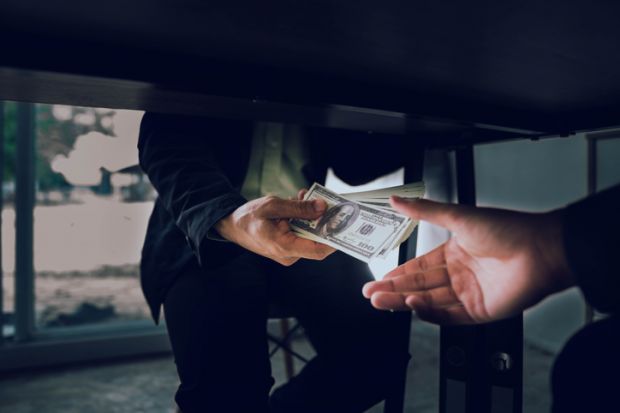 A man passes hundred-dollar bills under the table, illustrating corruption