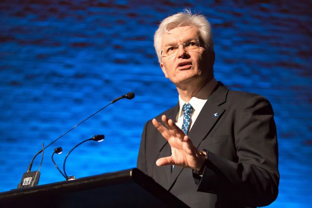 A man with grey hair and wearing a suit speaks at a podium 