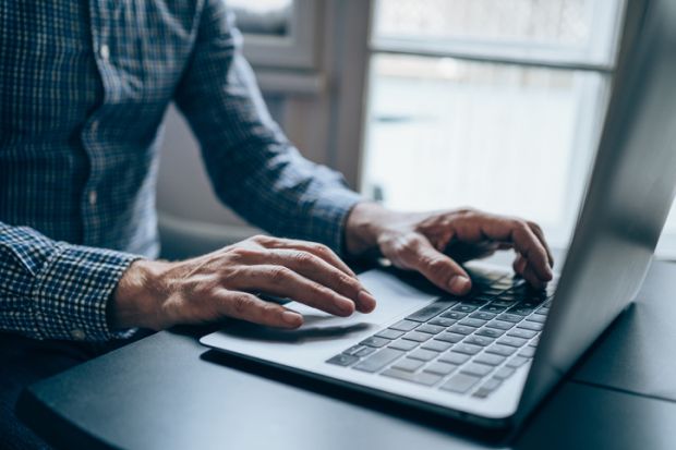 A man wearing a blue chequered shirt, whose face is not visible, typing on a laptop at a desk