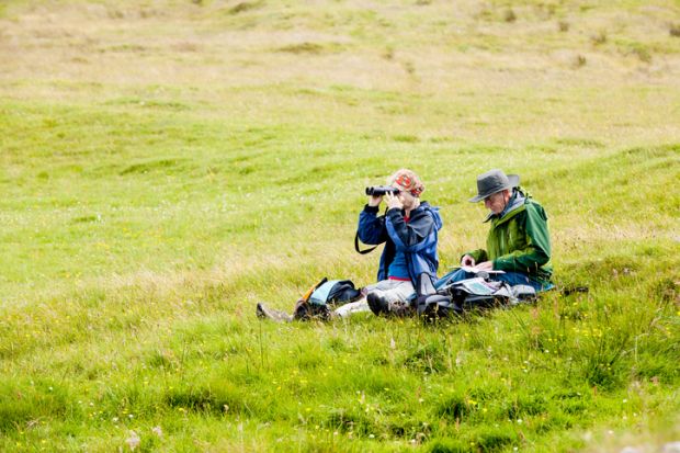 Two senior adults, a man and a woman, sit on the ground in a green meadow in Skye, Scotland