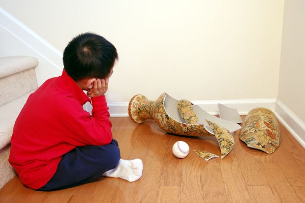 A young boy contemplates a smashed vase, with a ball sitting next to it