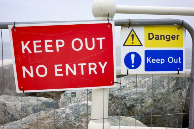 Red and blue 'Keep Out' signs on a fence by the sea, warning of danger ahead.