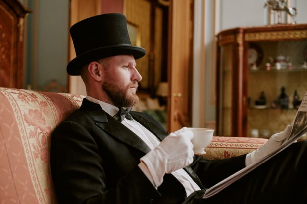 Man wearing formal suit and top hat sitting on sofa reading newspaper