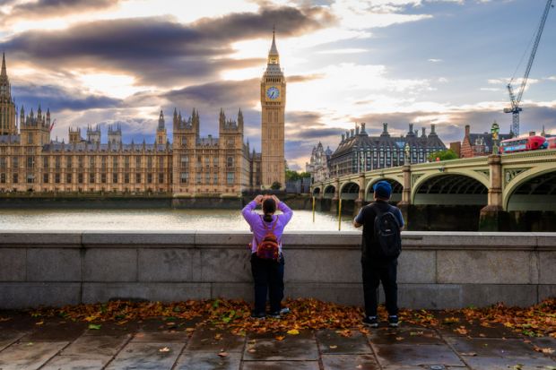 Rear view of a tourist couple taking photos of Big Ben and the Houses of Parliament from the south side of Westminster Bridge on the Thames embankment.