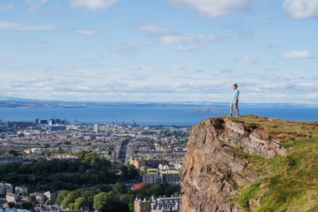 Standing on a rocky precipice, a person overlooks urban scenery and the ocean, under a bright sky