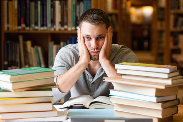 In a library, a student behind a pile of books clasps his face in his hands