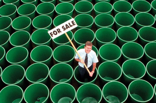 Man standing in rows of green plastic water butts, holding 'For sale' sign