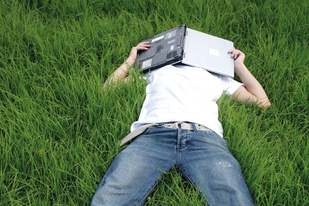 A man lying in the grass covering his face with a laptop