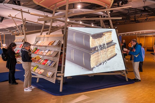 Visitors looking at books at a book fair, with a large image of water pouring out of a book. To illustrate that a number of institutions are predicted to drop deals with main scholarly imprints due to being to costly.
