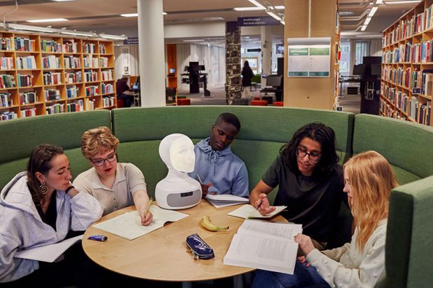 Students in a library with a robot head on the table. To illustrate AI risk to university jobs.