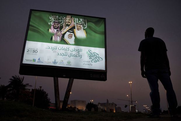 A pedestrian pauses near an illuminated sign showing portraits of the Saudi Arabian Royal family advertising Vision 2030 in Dhahran, Saudi Arabia. A pedestrian pauses near an illuminated sign showing portraits of the Saudi Arabian Royal family advertising Vision 2030 in Dhahran, Saudi Arabia.