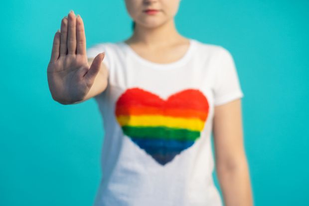 Stop gay discrimination. LGBT rights. Homophobia prevention. Unrecognizable woman with refusal hand gesture in t-shirt with rainbow flag heart on blue background.