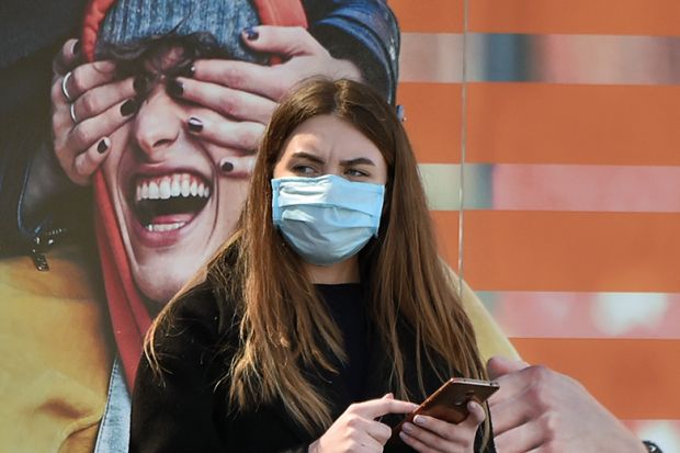 A woman wearing a face mask stands by an advertising placard showing someone having their eyes covered, April 21, 2020
