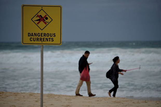 Visitors walk on Bondi beach next to a warning sign for dangerous sea currents on a stormy day in Sydney