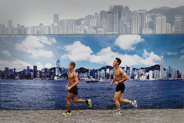 Two men jog past a billboard featuring photos of the city skyline with a clear sky on a cloudy day in Hong Kong