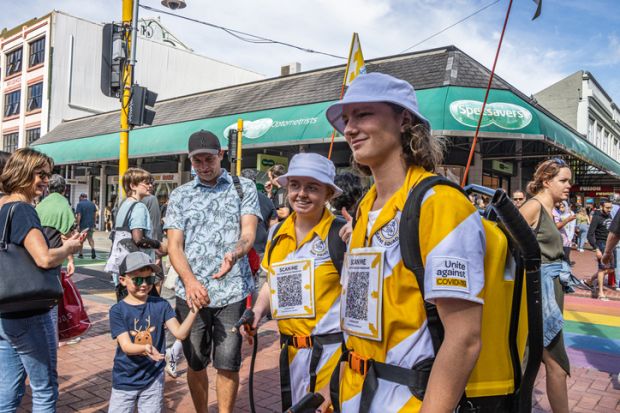 Two young ambassadors, armed with hand sanitiser pumps and scan codes for tracking whereabouts to assist with COVID-19 monitoring and tracking pose at the CubaDupa Street Festival in Wellington, while passersby take photos or wait for hand sanitiser.