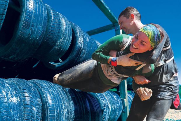 Man pulling lady out of tyres during an obstacle course race as a metaphor for Partners’ financial support is holding female academics back